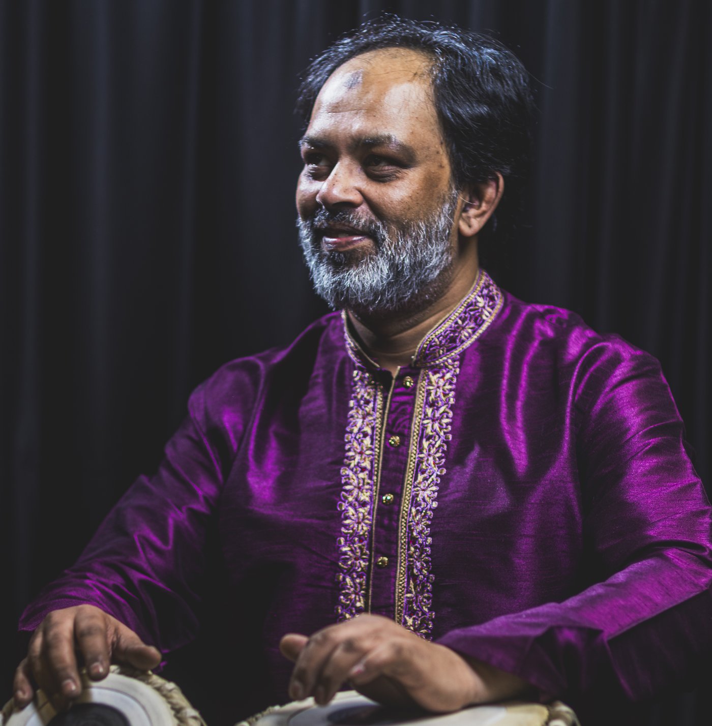 Ustad Shahbaz Hussain smiling as he plays tabla, in a purple embroidered kurta