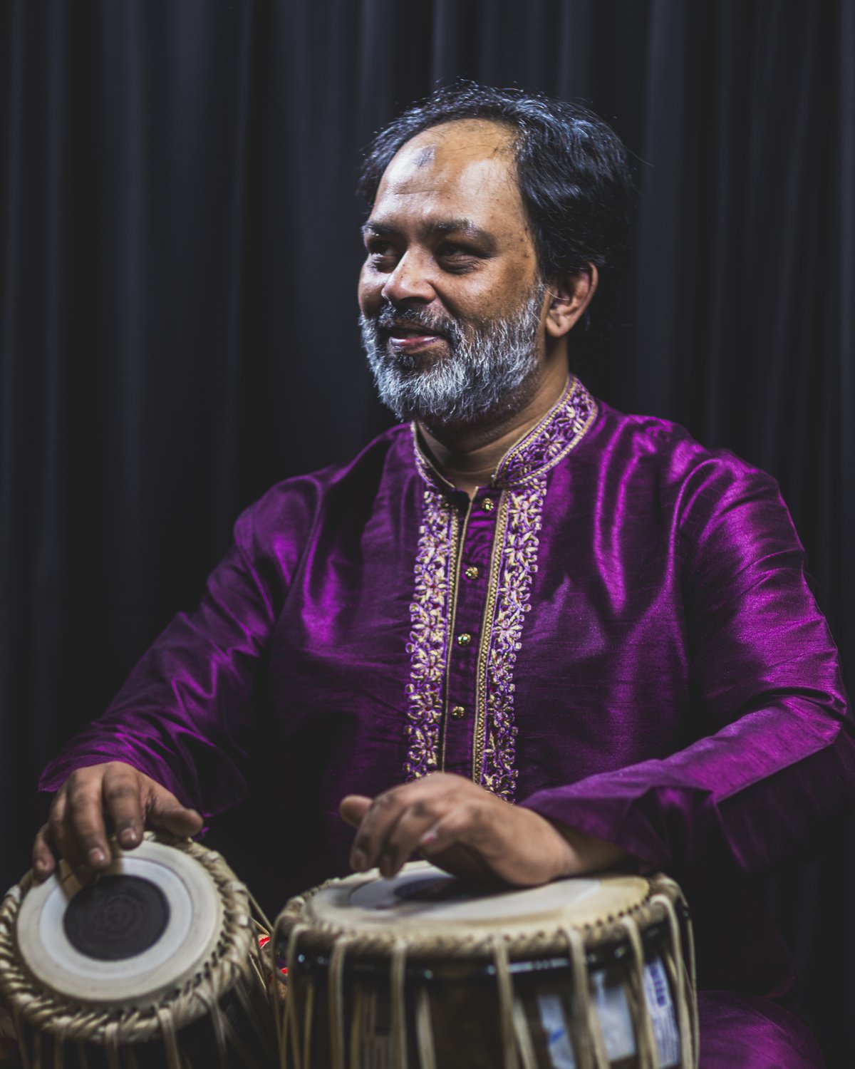 Portrait of Ustad Shahbaz Hussain in a purple embroidered kurta, hands on his tabla